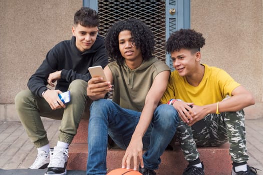 three happy multiethnic teen friends sharing mobile phone sitting together with on street, concept of youth and technology of communication