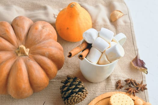 A cozy autumn setup featuring pumpkins, marshmallows in a cup, cinnamon sticks, and fall decorations.