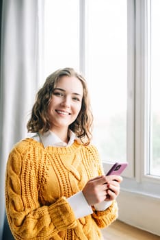 Smiling young woman using her smartphone at home, enjoying online shopping, messaging, and browsing. Capturing the essence of modern lifestyle and technology, she stands in a cozy indoor setting.
