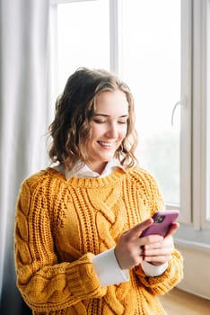 A cheerful young woman smiles while using her smartphone indoors at home. She browses online stores, texts messages, and downloads apps, portraying modern digital lifestyle and convenience.