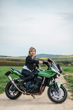 Woman in Leather Jacket on Green Motorcycle. A woman sits confidently on a green motorcycle parked on a dirt road, dressed in a leather jacket with red lipstick. Perfect for themes of adventure style.