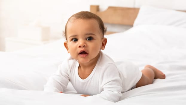 Cute African American Baby Infant In White Longsleeve Coverall Lying On Stomach In Bed Looking At Camera. Sweet Toddler Boy Posing Crawling In Bedroom At Home. Child Care, Clothes For Babies