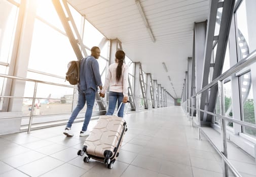 Travelling Together. Romantic African American Couple With Luggage Walking In Airport Terminal, Young Affectionate Black Spouses Holding Hands And Going To Flight Gate, Rear View With Free Space