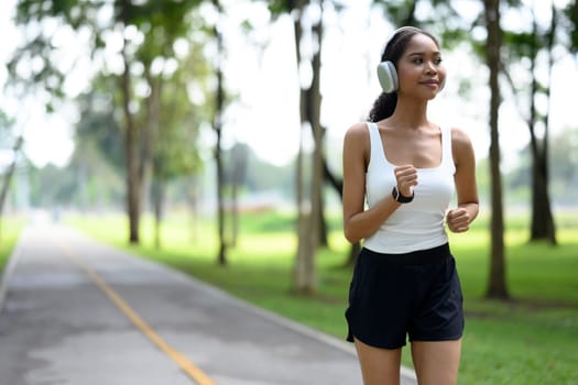Pleased sportswoman wearing headphones and a smartwatch jogging through the park.