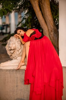 A woman in a red dress is sitting on a stone lion statue. The statue is white and has a lion's head. The woman is wearing a black belt and is looking at the camera. The scene has a calm