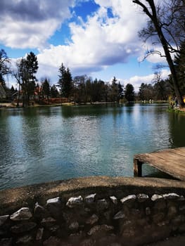 A calm lake reflects the cloudy sky, surrounded by trees and distant buildings, creating a peaceful natural environment perfect for relaxation.