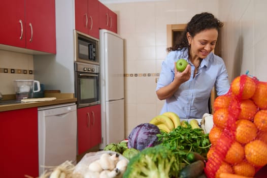A cheerful woman stands in a bright kitchen filled with fresh fruits and vegetables. She's selecting an apple, showcasing a healthy lifestyle and an abundance of nutritious food choices.
