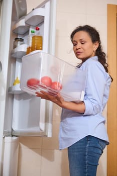 A woman stands in her kitchen, organizing her refrigerator with fresh red tomatoes. She wears a casual shirt and jeans, focusing on keeping her produce fresh and well-arranged.