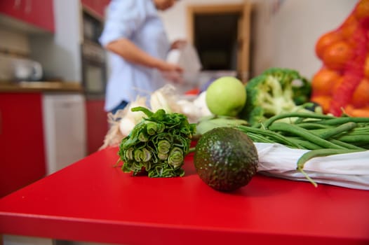 A close-up of vibrant vegetables and fruits on a red kitchen table, including an avocado, green beans, and broccoli, with a person preparing food in the background.