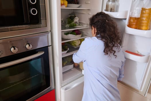 A person in a casual shirt organizing groceries in a refrigerator. The kitchen has a modern oven and fresh produce, suggesting home organization and healthy lifestyle.