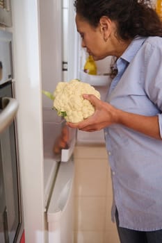 A woman holding a cauliflower searches inside a refrigerator. The scene conveys ideas of healthy cooking and fresh food choices. The open fridge indicates meal preparation at home.