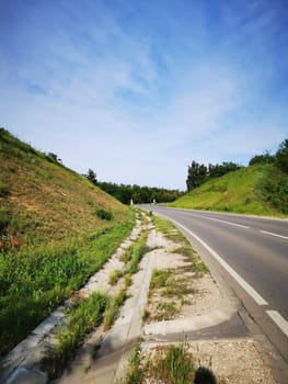 A tranquil country road curves gently between lush grassy slopes, with a backdrop of trees and a blue sky dotted with clouds during summer.