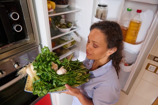 A woman stands in her kitchen holding a variety of fresh green vegetables near an open refrigerator. The scene conveys an emphasis on healthy eating, freshness, and home cooking.