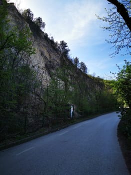 A tranquil rural road winds through lush greenery, with a steep hillside rising dramatically on one side under a clear blue sky, suggesting early spring in a serene landscape.