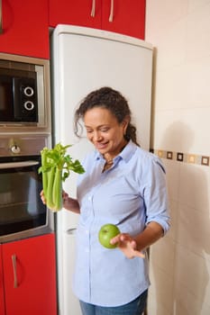 A woman stands in a kitchen holding green celery and an apple, embracing a healthy lifestyle. The modern kitchen with red cabinets adds a vibrant backdrop, reflecting health and wellness.