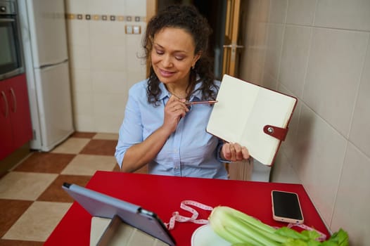 A woman in a kitchen video conferencing, holding a notebook. A tablet, tape measure, and vegetables are on the table. The scene conveys learning, planning, or remote work in a cozy setting.