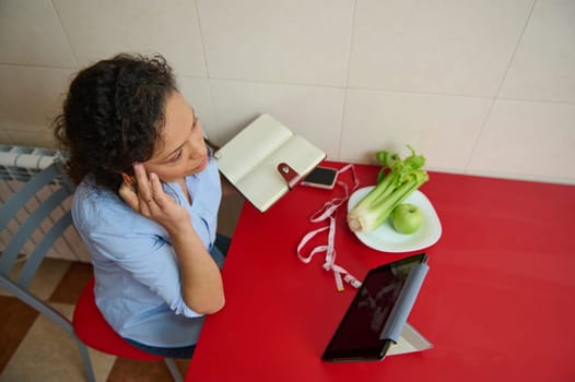 A woman sits at a red table, using her tablet to plan a healthy diet. Celery, an apple, and a measuring tape represent wellness and nutrition. She is focused and thoughtful on her goals.