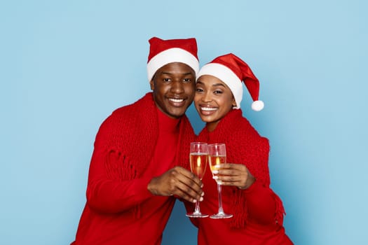 A happy black couple dressed in red holiday sweaters and Santa hats toast with champagne glasses against a light blue backdrop, capturing the spirit of Christmas celebration and togetherness.