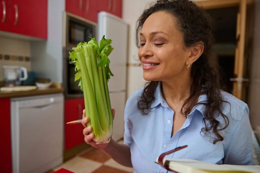 A smiling woman holds a fresh bunch of celery in her kitchen, embracing a healthy lifestyle. The bright kitchen setting enhances the feeling of freshness and positivity.