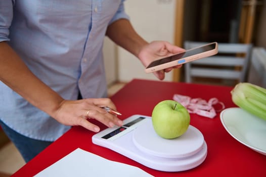 A person uses a smartphone app to weigh a green apple on a digital kitchen scale, emphasizing health monitoring and diet management. Celery and a measuring tape are visible in the background.