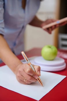 A person takes notes with a pen, while an apple and a digital tablet are nearby on a red table. This scene conveys focus, learning, and healthy lifestyle.