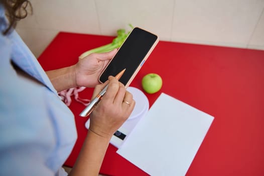 A person holding a smartphone and pen, recording health or diet-related data on a red counter beside a green apple. Organized planning for a balanced lifestyle is represented.