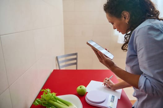A woman uses her smartphone to record dietary information at a table. Celery, an apple, and a scale are present, emphasizing a healthy lifestyle and wellness focus.