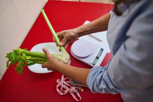 Person holding fresh celery stalks over a red surface with a digital scale and measuring tape. Focus on healthy eating, meal preparation, and accurate portion control.