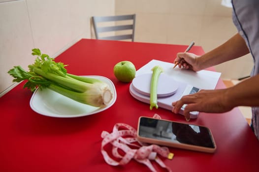 A person weighs a celery stalk on a kitchen scale, writing notes on a red table with a green apple, measuring tape, and phone nearby, symbolizing a healthy lifestyle and diet planning.