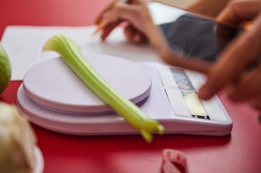 A fresh celery stalk rests on a digital scale as hands hold a smartphone. The image emphasizes healthy eating and meal planning with a focus on precision and nutrition.