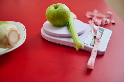 A fresh green apple and a celery stick rest on a digital kitchen scale on a red table. A measuring tape nearby suggests a focus on health or diet planning, embodying fitness goals.