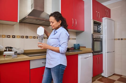 A woman in a casual outfit is standing in a modern red kitchen, pouring filtered water into a glass. The scene conveys a sense of routine and domestic life in a cozy home setting.