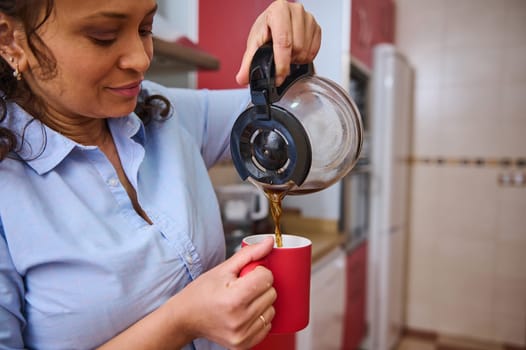 A woman in a light blue shirt pours steaming coffee into a red mug with a peaceful smile. The warm kitchen backdrop adds a homely feel, capturing a moment of tranquility.