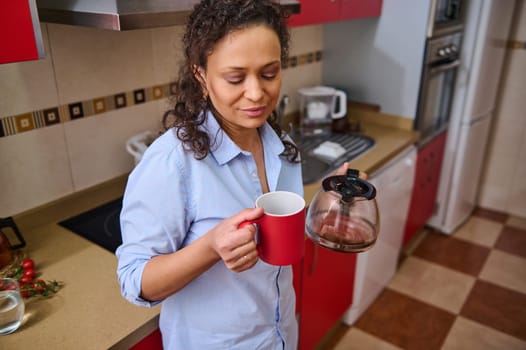 A woman stands in a modern kitchen, enjoying a fresh cup of coffee. She appears relaxed and content, taking a moment from her daily routine. Red cabinet and countertop add to the cozy ambiance.