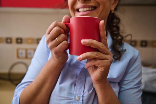 A smiling woman holds a red mug in a cozy setting, enjoying a warm drink. The image captures a moment of relaxation, warmth, and contentment, perfect for themes of home and comfort.