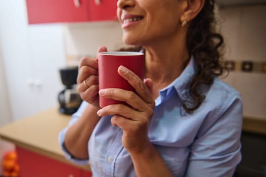 A woman holds a red mug, enjoying a warm drink in a cozy kitchen. Her relaxed expression suggests comfort and calm, creating a peaceful, homey atmosphere.