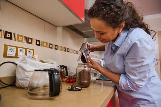 A woman in a blue shirt pours coffee grounds into a jar in her kitchen, surrounded by kitchen items. She smiles, enjoying the simple task in her cozy home environment.