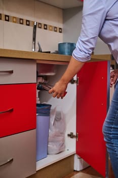 A person is repairing a kitchen sink with tools, with the red cabinet door open. The scene emphasizes home maintenance and plumbing work in a modern kitchen setting.