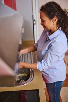 Side view of a woman washing dishes in a stylish kitchen. She's wearing a casual blue shirt, focused on cleaning. The setting is warm and homely, emphasizing daily routine and responsibility.