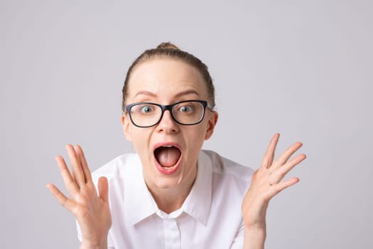 A surprised woman with glasses expressing shock or excitement, hands raised in a dramatic gesture. She has a neutral background and is wearing a white shirt.