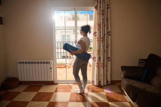 A woman stands in a bright room, holding a blue yoga mat and looking outside. The sunlight streams in through the glass doors, creating a peaceful atmosphere ideal for home fitness.