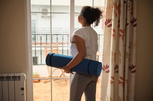 A woman stands inside her home holding a blue yoga mat, gazing out the window. The scene conveys relaxation and preparation for a workout or meditation session.