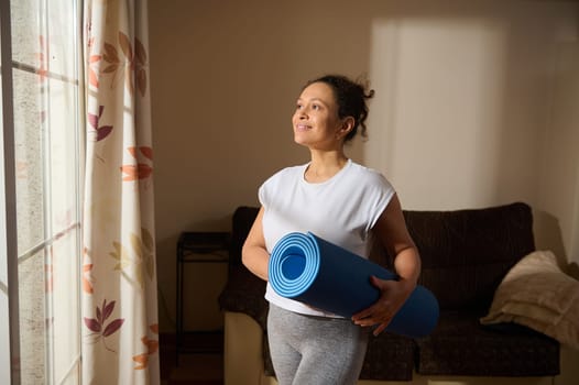 A woman stands in a living room holding a yoga mat, basking in warm sunlight. She exudes a sense of calm and readiness, embodying a healthy and mindful lifestyle.