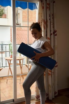 A woman stands near a sunny window, holding a blue yoga mat, with a peaceful expression. The setting is a cozy home with a view outside, capturing a moment of calm and readiness for exercise.