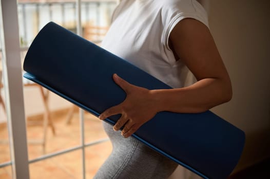 A woman carrying a blue rolled yoga mat stands in a bright room, ready for exercise. The scene conveys a sense of calm and readiness, showcasing a healthy and active lifestyle.