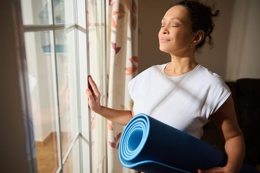 A serene woman stands by a window, eyes closed, holding a yoga mat. She basks in the sunlight, embodying peace and relaxation. It's a moment of mindfulness and tranquility at home.