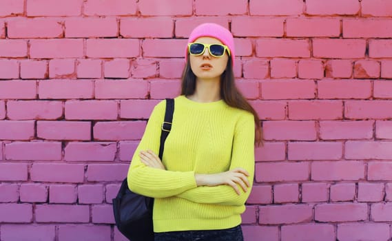 Stylish young teenager girl with backpack posing in vivid colorful clothes, yellow sweater, hat, glasses with backpack on city street on pink brick wall background