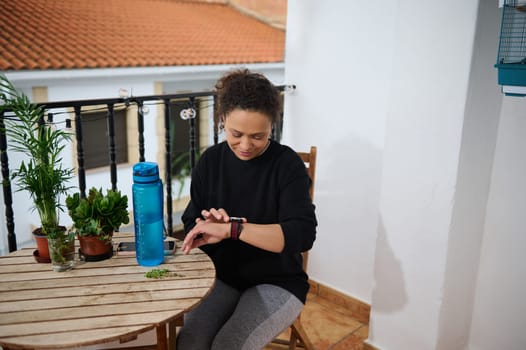 A woman seated on a balcony looks at her smartwatch, suggesting a break or checking progress during a home workout session. The scene includes a water bottle and plants, conveying relaxation.