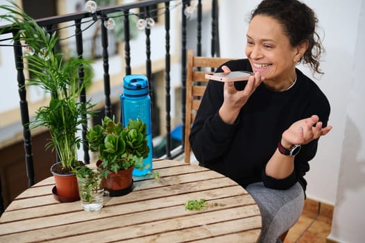 A cheerful woman engages in a phone conversation on a cozy terrace, surrounded by plants and a water bottle. Her genuine smile reflects happiness and connection in a relaxing outdoor setting.