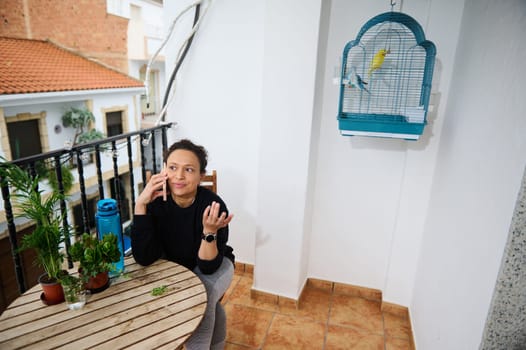 A woman relaxes on her balcony, chatting on the phone. Surrounded by potted plants and a colorful birdcage, she seems content in this cozy outdoor space, reflecting a harmonious lifestyle.